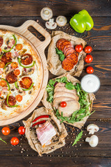 Large meat pizza with salami, bacon, mushrooms, tomatoes and paprika on a round cutting board on a dark wooden background. Ingredients.