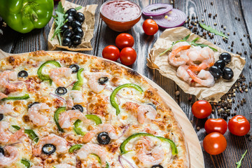 Big pizza with seafood, shrimps and black olives on a round cutting board on a dark wooden background. Food Ingredients. Macro