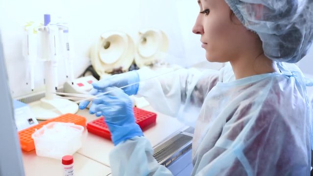 A young female scientist works in a sterile genetic or bacteriological laboratory with a dispenser and reagents in a medical gown, gloves and a doctor's cap.