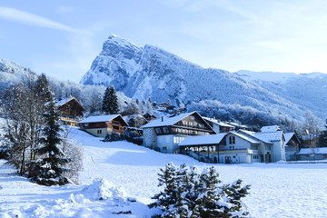 Fototapeta premium Alpine chalets covered with snow on slopes by a big rock and pine tree forest in French skiing resort .
