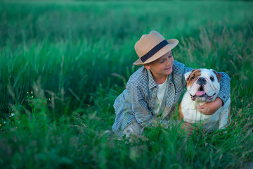 Little boy sitting standing with his english bull dog on the meadow of green rye. Handsom kid posing with best friend wearing stylish clothes and rustic retro hat