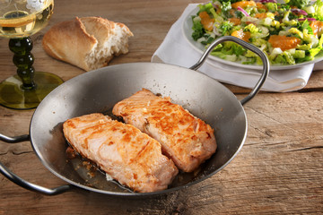fried salmon fillet in a pan on a rustic wooden table,  salad bread and wine in the background