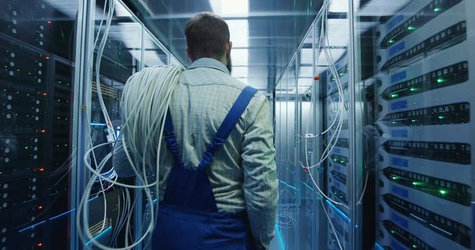 Wide movement stabilized shot of two people working in a data center carrying cable to repair rows of server racks and discuss their work