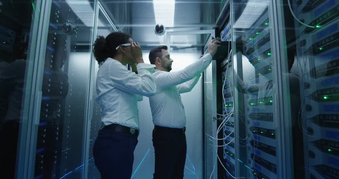 Medium shot of two diverse employees working in a data center server room taking networking equipment out for repair and maintenance