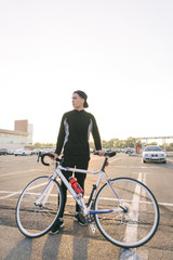 Fototapeta premium Young man is a cyclist with dark sportswear and a cap with a bicycle on the backdrop of the city landscape and looking to the side. Cycling Sports Concept.