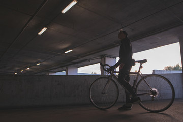 An evening photo of a young cyclist standing under the bridge with a bicycle. Background with a cyclist in dark colors.
