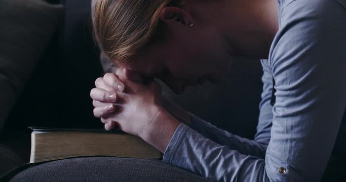 A slow cinematic dolly shot of a young woman with a Bible praying at home.