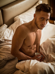 Totally naked sexy young man with muscular body on bed with mug or cup in hand with coffee or tea