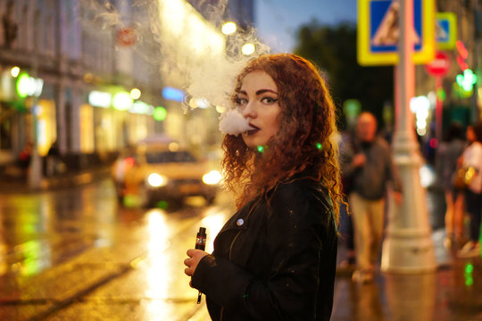 Young attractive red-haired girl smokes electronic cigarette. She is in leather jacket standing on road in street of night city. Wet asphalt after rain.