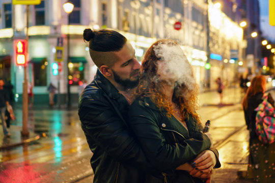 Couple in love on date. Boyfriend hugs girl from behind. She smokes an electronic cigarette. They walk streets of night city. Wet asphalt after rain.