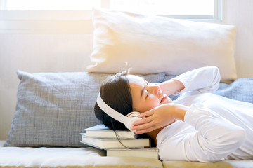 Asian charming beautiful smiling women listens music or relax sound from her headphones and put her head on stack of books in the living room on sofa at the morning time.