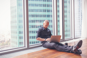Young attractive stylish bald man in a business suit sitting on the floor with a laptop in his office in a skyscraper, business, finance, success