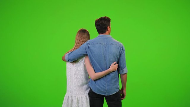An in-love couple is looking to the horizon, smiling and hugging, over a green screen, from the back.