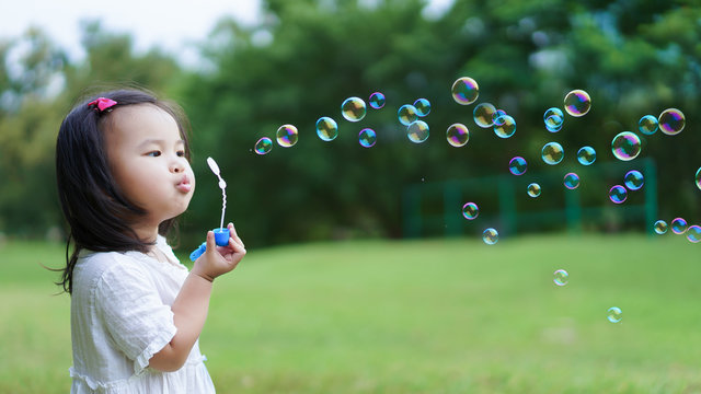 Asian cute little girl blowing to make many bubbles in public garden at holiday or vacation. Playtime Concept.