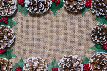 Christmas flat-lay frame of pinecones, holly leaves, and red berries on rustic fabric background