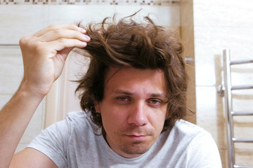 Sleepy shaggy young man looks at the mirror in bathroom in the morning.