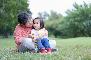 Asian cute happy little granddaughter sit on grandmother's lap and enjoy to play with her at public park or garden in vacation or holiday. Family Concept.