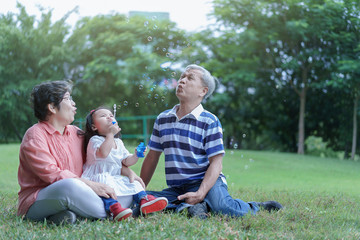 Asian attractive happy grandfather help to blows the bubbles from granddaughter made them and attractive happy grandmother look at them in the public garden at vacation or holiday. Family Concept.