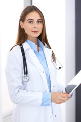 Doctor woman using tablet computer while standing straight near window in hospital. Happy physician at work. Medicine and health care concept