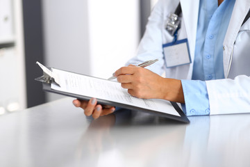 Unknown doctor woman filling up medical form while sitting at the desk in hospital office, close-up of hands. Physician at work. Medicine and health care concept