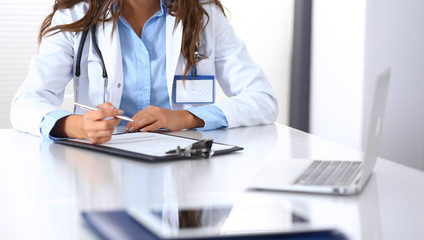 Unknown doctor woman filling up medical form while sitting at the desk in hospital office. Physician at work. Medicine and health care concept