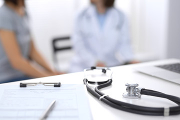 Stethoscope, clipboard with medical form lying on hospital reception desk with laptop computer and busy doctor and patient communicating at the background. Medical tools at doctor working table