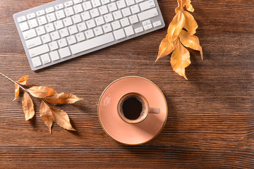 Cup of aromatic coffee, keyboard and autumn leaves on wooden background, top view