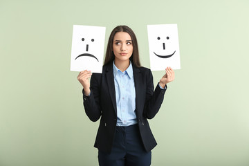 Young woman holding sheets of paper with drawn emoticons on light background