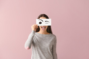 Emotional young woman hiding face behind sheet of paper with drawn eyes on color background - obrazy, fototapety, plakaty