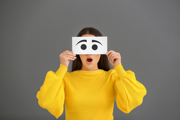Emotional young woman hiding face behind sheet of paper with drawn eyes on grey background