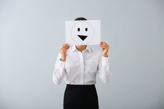 Young woman hiding face behind sheet of paper with drawn emoticon on light background