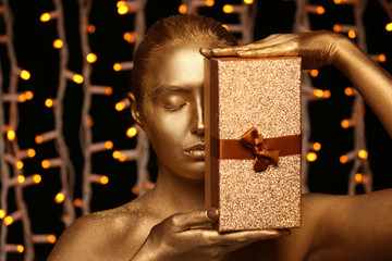 Beautiful young woman with golden paint on her body and gift box against defocused lights