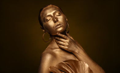 Fototapeta na wymiar Beautiful young woman with golden paint on her body and tropical leaf against dark background