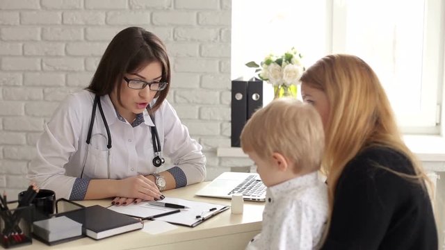 Kind and sweet woman doctor talking with a little boy in a children's clinic. Mom with a little son at a pediatrician, the doctor is very nice talking with the baby. Little boy is afraid of the doctor
