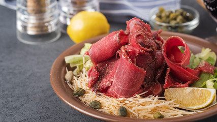 Cold snack with raw frozen meat stroganina with cheese, capers and lemon. Sliced frozen raw meat on a plate.