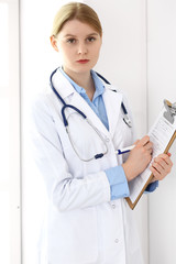 Doctor woman  using clipboard with medical record form while  standing near window in clinic or hospital. Medicine and healthcare concept. Physician at work