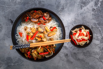 Traditional Asian food - rice noodles with seafood, salad, red pepper and fried mushrooms are on the side table. Top view
