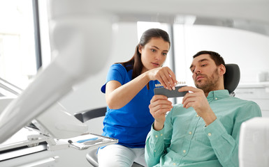 medicine, dentistry and healthcare concept - female dentist with tooth color samples choosing shade for male patient teeth at dental clinic