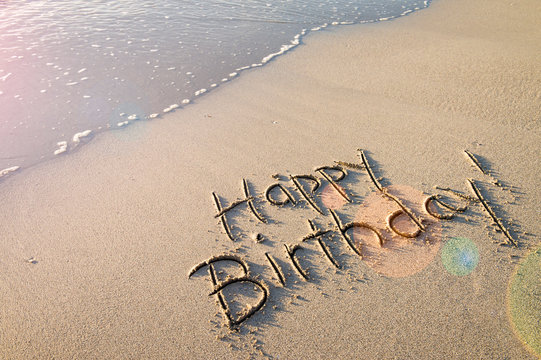 Happy Birthday! message handwritten in smooth sand with festive lens flare on the shore of an empty beach