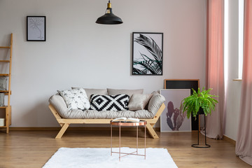 Bright couch with pillows in real photo of white sitting room interior with posters, black lamp and pastel pink drapes