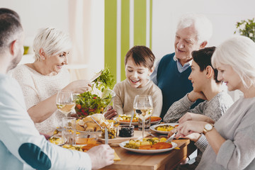 Smiling boy sitting on grandfather's knees during family dinner with parents and grandmother