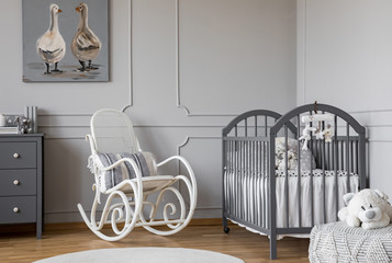 White rocking chair with pillow next to wooden cradle in elegant baby room with duck's poster on the wall