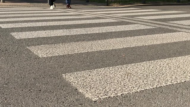 Few pedestrians cross over small road by zebra crossing at sunny day. Pedestrian foot thwart the road by street crosswalk in city. People step across a road by panda crossing on green light