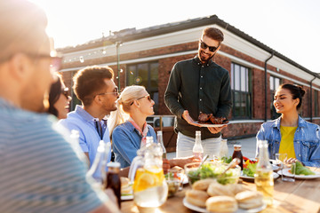 leisure and people concept - happy party host offering meat to his friends at barbecue party on rooftop in summer