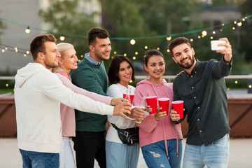 leisure and people concept - happy friends with drinks in party cups taking selfie by smartphone on rooftop in summer