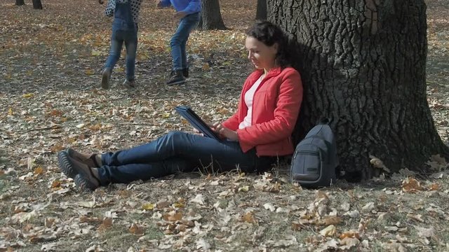 Family in nature in the fall. Mother with tablet, daughters playing together in the autumn park.
