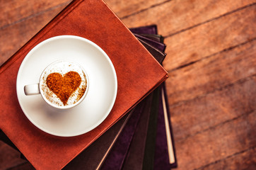 white cup of coffee with cream heart shape symbol on books on wooden background
