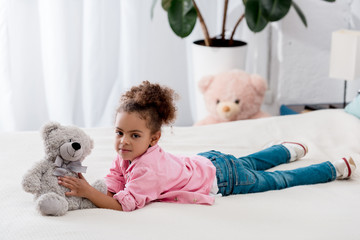 Curly african american kid lying on the bed with her teddy bear