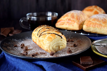 Puff pastry bun decorated with chocolate on a dark plate sprinkled with sugar powder