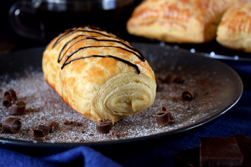 Puff pastry bun decorated with chocolate on a dark plate sprinkled with sugar powder